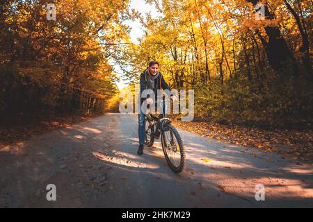 Ein Mann auf dem Fahrrad fährt am Abend bei Sonnenuntergang entlang der Straße im Herbstwald Stockfoto