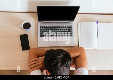 Der junge Mann schläft auf dem Tisch vor dem Computer, der sich auf seine Arme stützt, mit einem Kaffee, einem Smartphone und einem Notebook. Konzept: Telearbeit oder havi Stockfoto