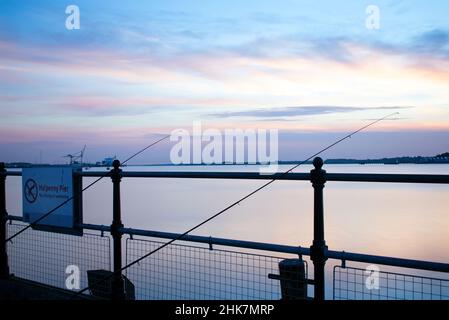 Zwei Angelruten stützen sich auf das Metallgeländer des historischen Ha'Penny Pier in Harwich, North Essex, Großbritannien. Der internationale Hafen von Harwich ist flussaufwärts zu sehen Stockfoto