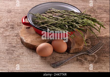 Roher wilder Spargelstrauß in einem alten roten emaillierten Topf auf einem Holztisch Stockfoto