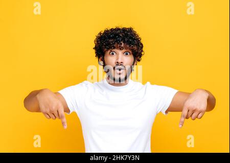 Positiver indischer oder arabischer glücklicher Kerl im weißen T-Shirt, staunend blickt auf die Kamera und zeigt die Finger nach unten, überraschender Gesichtsausdruck, steht auf isoliertem orangefarbenem Hintergrund Stockfoto