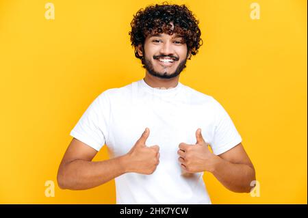 Fröhlicher, fröhlicher, junger, erwachsener inder oder araber, gekleidet in ein einfaches weißes T-Shirt, zeigt Daumen nach oben mit zwei Händen, blickt auf die Kamera, steht auf einem isolierten orangefarbenen Hintergrund und lächelt freundlich Stockfoto