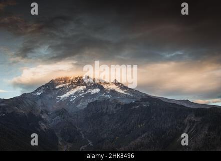 Eine linsenförmige Wolke umschließt den schneebedeckten Berg Hoot bei einem wunderschönen Sonnenuntergang im Herbst. Mit einem meist blauen Himmel gibt es eine Bergkette von Gipfeln Stockfoto