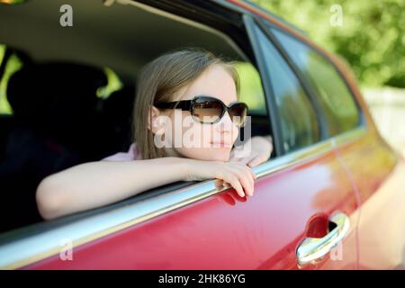Lustiges junges Mädchen, das ihren Kopf aus dem Autofenster ragt und sich auf eine Roadtrip oder Reise freut. Familienauto reisen mit Kindern. Stockfoto