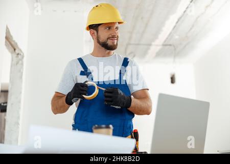 Selbstbewusster Handwerker, Elektriker lächelt beiseite und hält Klebeband für die Installation einer neuen Steckdose in der Wohnung Stockfoto