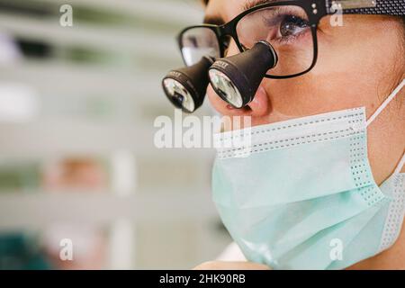 Junge Ärztin, die bei ihrer Arbeit in der Zahnarztpraxis ein Dentalluppenfernglas und eine medizinische Maske trägt. Stockfoto