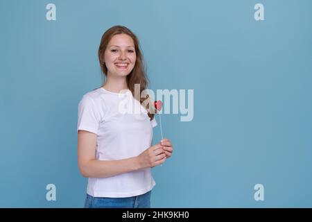 Valentinstag, Mädchen auf einem blauen Hintergrund hält Herz. Blondine lächelt schön, lacht liebt. März 8, Geschenke für Mädchen. Rotes Herz. Kaukasische junge Frau in weißem T-Shirt Stockfoto