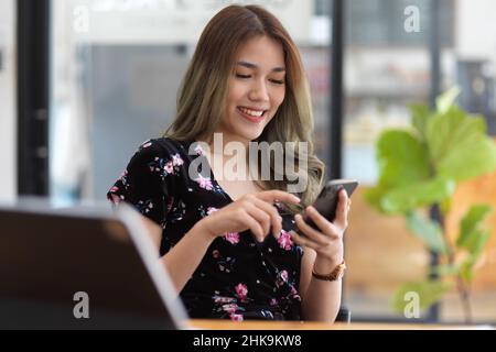 Charmante asiatische tausendjährige Frau in Casual Outfit entspannen in gemütlichen Café und mit ihrem Mobiltelefon. Text, Nachricht, Social Media, Anwendung Stockfoto