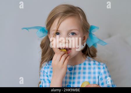 Portrait glückliches kleines kaukasisches Mädchen, das knusprige Kartoffelchips auf grauem Hintergrund in einem blauen Kleid isst. Kind liebt es, Konzept zu essen. Cheat Mahlzeit und Fast Food. Panorama, leerer Raum Stockfoto