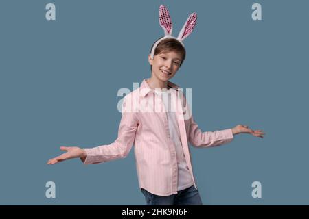 Foto angenehmer junger Mann mit lustig kindlicher Stimmung, gestikuliert mit den Händen und schaut in die Kamera, mit rosa Ohren eines osterhasen mit einem Verband an den Ohren in einem lässigen rosa Hemd isoliert auf blau Stockfoto