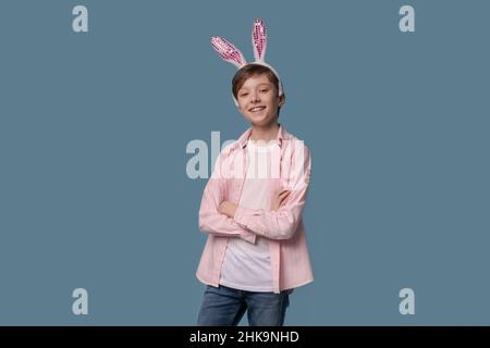 Foto angenehmer junger Mann mit lustig kindlicher Stimmung, gestikuliert mit den Händen und schaut in die Kamera, mit rosa Ohren eines osterhasen mit einem Verband an den Ohren in einem lässigen rosa Hemd isoliert auf blau Stockfoto