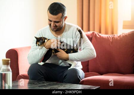 Nahaufnahme eines Mannes, der mit einer Hauskatze und einem gemütlichen roten Sofa jubelt und genießt. Stockfoto