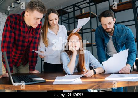 Erschöpft erschöpfte junge Geschäftsfrau unter Stress im Büro am Schreibtisch sitzend mit multiethnischen Kollegen, die Papierdokumente in ihrer Nähe schüttelten. Stockfoto