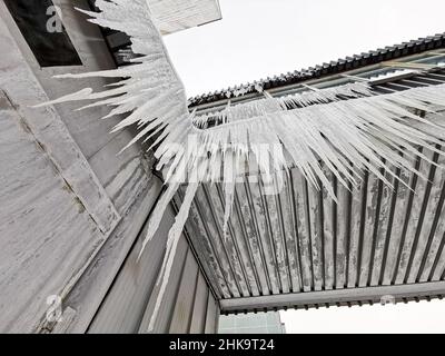 Viele lange durchsichtige Eiszapfen hängen vom Dach des Gebäudes. Ansicht von unten. Scharfe Kanten von glänzenden Eiszapfen in Nahaufnahme. Stockfoto