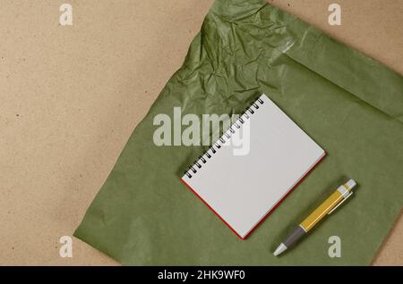 Ein offenes Notizbuch mit leeren weißen Blättern und einem gelben Stift auf einer mehrfarbigen Oberfläche. Grünes zerknittertes Papier vor beigefarbenem Hintergrund. Draufsicht. S Stockfoto