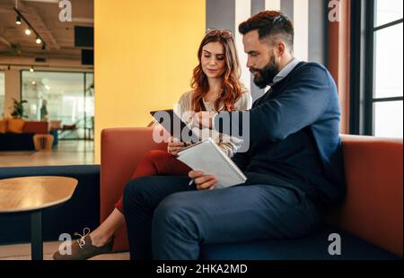 Zwei fokussierte Geschäftsleute, die in einer Bürolobby ein digitales Tablet nutzen. Zwei moderne Geschäftsleute, die in einem Co-Working-Space eine wichtige Diskussion führen. Stockfoto