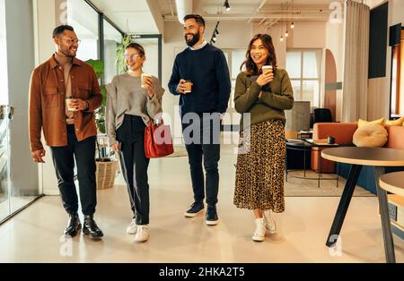 Verschiedene Geschäftsleute, die morgens durch ein Büro laufen. Ein Team glücklicher Geschäftsleute, die lächeln, während sie Kaffeetassen halten. Unternehmergruppe Stockfoto