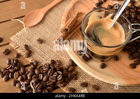 Glas Tasse heißen Kaffee und auf Holzsockel und Kaffeebohnen auf Sackleinen. Erhöhte Ansicht. Horizontale Zusammensetzung. Stockfoto