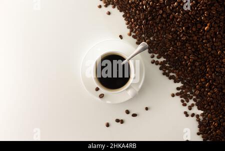 Weiße Tasse Kaffee auf der Untertasse auf dem weißen Tisch und Stapel Kaffeebohnen in einer Ecke. Draufsicht. Horizontale Zusammensetzung. Stockfoto