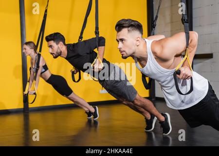 Junger Sportler, der im Fitnessstudio an einem Federband neben verschwommenen multiethnischen Freunden zieht Stockfoto