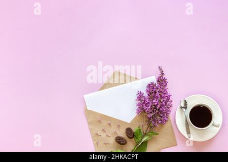 Zweig Flieder auf Bastelumschlag mit weißer leerer leerer Karte für Text und Tasse Kaffee, rosa Hintergrund. Grußkarte Flat Lay Mock Up Concept Hell Stockfoto