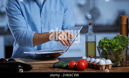 Beschnittene Ansicht eines Mannes, der ein Kochbuch hält und auf frisches Gemüse auf dem Tisch zeigt Stockfoto