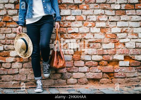 Die Frau in einer Jeansjacke mit Strohhut steht auf einer Ziegelwand. Trendige Frau in legerer Kleidung. Modekonzept Stockfoto