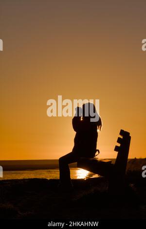 Silhouette einer jungen Frau mit Fernglas Vogelbeobachtung auf Einer Bank im Sunset Keyhaven UK Stockfoto