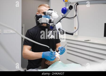 Ein professioneller junger, männlicher Zahnarzt verwendet ein Mikroskop, um eine weibliche Patientin zu behandeln. Moderne Technologien in der Zahnmedizin Stockfoto