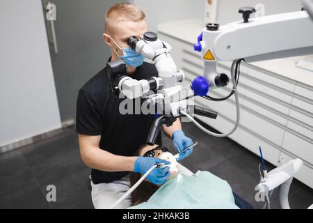 Ein professioneller junger, männlicher Zahnarzt verwendet ein Mikroskop, um eine weibliche Patientin zu behandeln. Moderne Technologien in der Zahnmedizin Stockfoto
