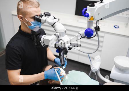 Ein professioneller junger, männlicher Zahnarzt verwendet ein Mikroskop, um eine weibliche Patientin zu behandeln. Moderne Technologien in der Zahnmedizin Stockfoto