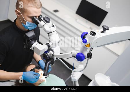 Ein professioneller junger, männlicher Zahnarzt verwendet ein Mikroskop, um eine weibliche Patientin zu behandeln. Moderne Technologien in der Zahnmedizin Stockfoto