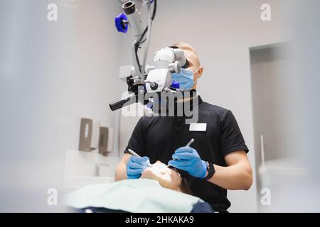 Ein professioneller junger, männlicher Zahnarzt verwendet ein Mikroskop, um eine weibliche Patientin zu behandeln. Moderne Technologien in der Zahnmedizin Stockfoto