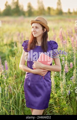 Frau im Sommerfeld mit Blumenstrauß - Lupinen, Hut und rosa Notizbuch. Nähe zur Natur, Selbsterkennungskonzept. Entspannen und Wohlfühlen Stockfoto