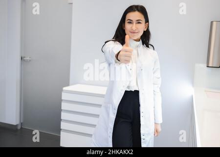 Positive junge Ärztin, die die Kamera anschaut und die Daumen nach oben zeigt Stockfoto
