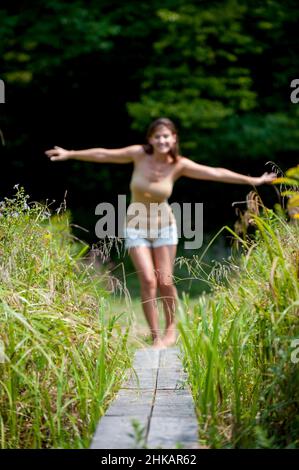 Junge Frau balanciert auf einem schmalen Holzsteg, die Arme sind ausgestreckt, um das Gleichgewicht zu unterstützen Stockfoto