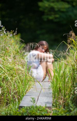 Die junge Frau saß auf einem schmalen Holzsteg, die Arme um ihre Beine waren von hohen Gräsern umgeben Stockfoto