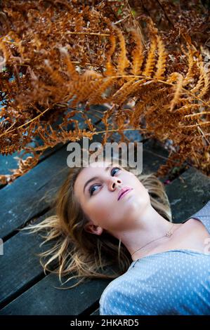 Junge Frau, die sich auf einer hölzernen Veranda niederlegt, umgeben von Farnen, mit dem Blick nach oben Stockfoto