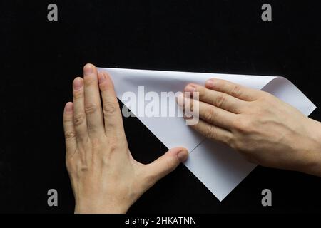 Frau Hände Falten rechteckigen Blatt Papier, um es quadratisch auf schwarzem Hintergrund zu machen Stockfoto