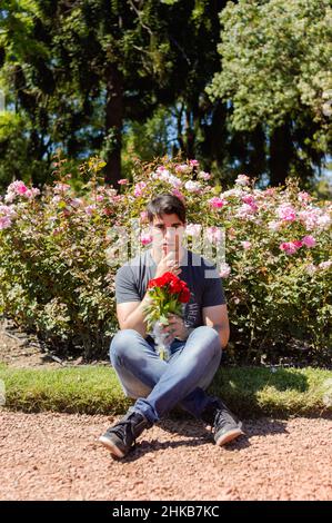 Junger kaukasischer lateinamerikanischer Mann, der in einem Rosengarten im Freien in einem Stadtpark sitzt, mit einem Blumenstrauß in der Hand und Blick auf die Kamera, mit fl Stockfoto