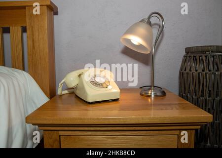 Ein altes cremefarbenes Telefon im Retro-Stil auf einem Nachttisch im Schlafzimmer mit einer Lampe, die auf das Telefon zeigt Stockfoto