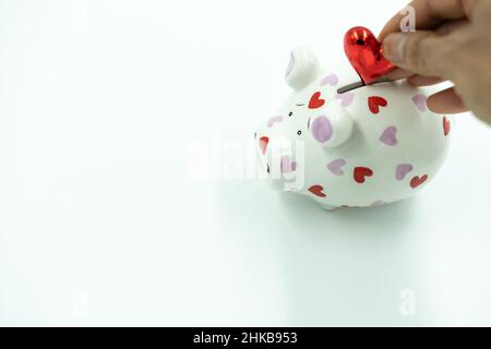 Hand aus Mischling Frau füllt weißen Sparschwein mit rotem Herz auf weißem Hintergrund. Stockfoto