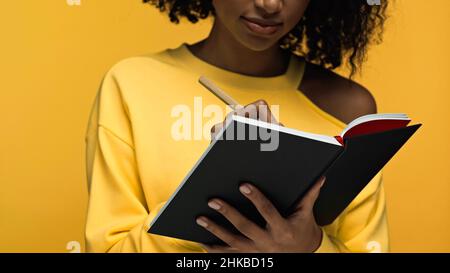 Beschnittene Ansicht einer jungen afroamerikanischen Frau, die in einem Notizbuch auf Gelb geschrieben hat Stockfoto