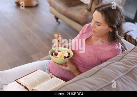 Kaukasische Schwangere, die einen Snack isst, während sie ein Buch liest, während sie zu Hause auf der Couch sitzt Stockfoto