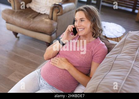 Kaukasische Schwangere berührt ihren Bauch und spricht auf dem Smartphone, während sie auf der Couch liegt Stockfoto