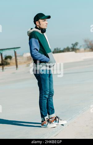 Ein Mann mittleren Alters, der moderne Freizeitkleidung und ein Paar Kopfhörer im Nacken trägt, steht auf der Promenade, neben dem Sand eines städtischen Strandes Stockfoto