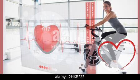 Digitales Composite-Bild von Herzformen und Pulsverlauf über kaukasische junge Frau beim Radfahren im Fitnessstudio Stockfoto