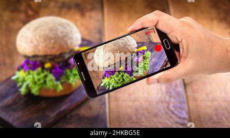 Männliche Hand hält Handy schwarz Smartphone macht Video von Lebensmitteln, Hamburger für Website oder Lieferung App gefilmt Stockfoto