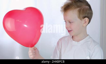 Valentinstag, Kinder. Ein süßer kleiner Junge am Valentinstag oder Muttertag lächelt, mit einem roten Ballon in den Händen, in Form eines Herzens, auf ein Whi Stockfoto