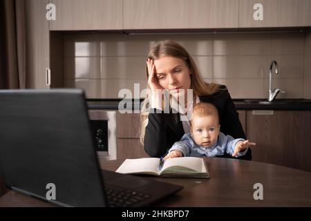 Stress beim Mutterschaftsurlaub. Eine junge Frau mit einem Baby in den Armen leidet unter Kopfschmerzen, während sie in einem Heimbüro an einem Laptop arbeitet, das hat eine kranke Mutter Stockfoto